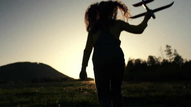 Silhouette of happy little girl child running with toy airplane on field in sunset light. Children play toy airplane on sky background. Preteen dreams of flying and becoming pilot. Slow motion
