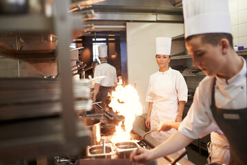 Cooking, chef and team in kitchen with fire on frying pan at restaurant for fine dining, service and training. Expert, culinary professional and group of people preparing dinner for food catering