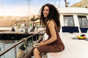 A cheerful woman enjoys reading a book on a yacht - relaxed lifestyle at a sunlit marina.