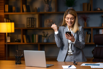 Cheerful  woman in gray  suit holding smartphone and celebrate victory triumph while standing in office room.