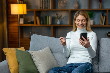 Smiling and excited adult woman checking her recent pregnancy test, sitting on gray couch at home