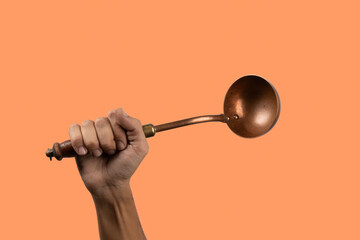 Black male hand holding a brass vintage kitchen ladle on orange background