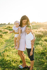 Fototapeta premium Portrait of daughter toddler and son hugging mother in nature on summer day. Mom hugs children standing in field at sunset. Concept of friendly family. Closeup. Mommy and kids spend time together.