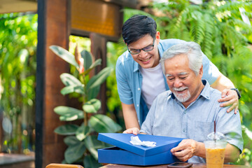 Asian man surprised elderly father with Birthday gift at outdoor cafe restaurant on summer holiday vacation. Family relationship, celebrating father's day and senior people health care concept.