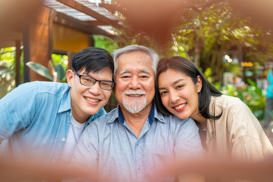 Happy Asian couple using mobile phone taking selfie with elderly father during having lunch eating food together at restaurant on summer vacation. Family relationship senior people health care concept