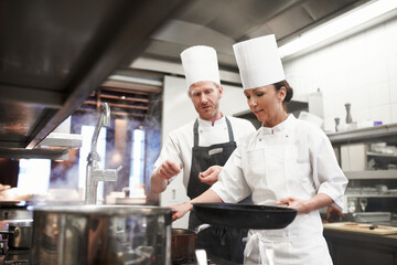 Chef, teamwork and kitchen or pan cooking for hospitality service as preparation, learning or catering. Man, woman and hat at stove for teaching skill for fine dining presentation, recipe or cuisine - Powered by Adobe