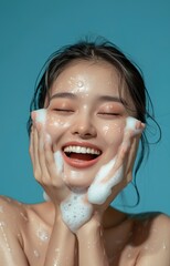 A young Asian woman washing her face with white foam, smiling and looking happy against a plain background. Skincare concept