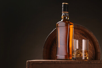 Whiskey with ice cubes in glass and bottle on wooden table near barrel against dark background,...