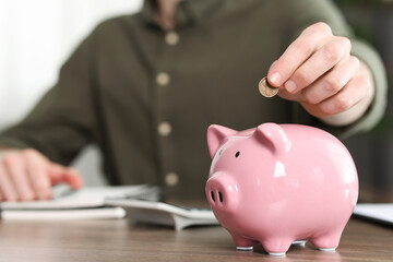 Financial savings. Man putting coin into piggy bank at wooden table, closeup
