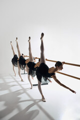 Four ballet dancers line up at barre, performing identical pose with outstretched leg, training against grey studio background. Concept of ballet, art, dance studio, classical style, youth
