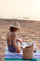 Young woman in straw hat setting on the beach striped towel with jute bag. Summer vacation lifestyle concept