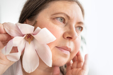 mature woman 55 years old with magnolia flower gazes into mirror, observing wrinkles, shifts in...