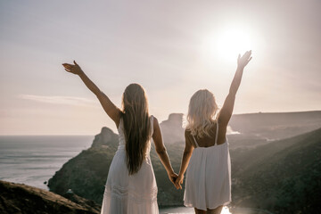 Two women are standing on a hill overlooking the ocean. They are holding hands and looking out at...