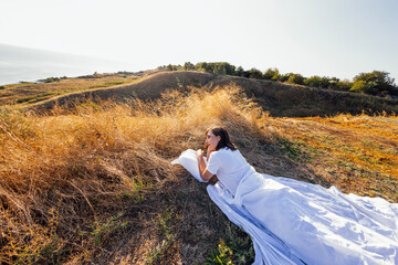 A young woman in white clothes lies on a white sheet and pillow in a field among dry grass. The girl relaxes and rests in nature.