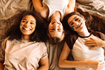 Lovely teenagers of different races apply cosmetic masks to their faces in the bedroom. Multiethnic friends are lying on the bed and having fun at home together.
