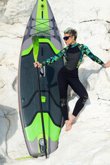 A beautiful sup surfer girl in a wetsuit on the ocean shore against a beautiful blue sky poses...