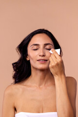 Studio portrait of an attractive young woman applying moisturizing cream to her face under her eyes on a beige isolated background.