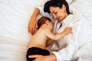 A cute baby sleeps with his attractive mother in a bed. A little boy falls asleep in the arms of his mother.