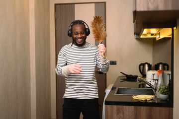 Cheerful man in headphone enjoying domestic chores, cleaning up his house and singing - obrazy, fototapety, plakaty