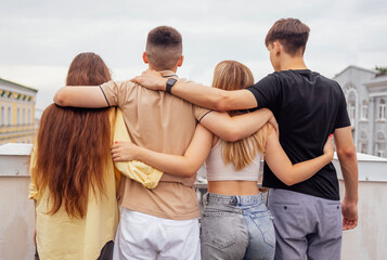Four teenagers hug and stand with their backs on the roof of the house. Friends have a great time...