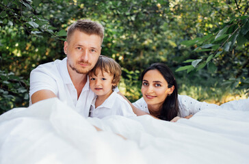 Portrait of happy cute family in the garden. Mom, dad and baby are posing and looking at the camera. - obrazy, fototapety, plakaty