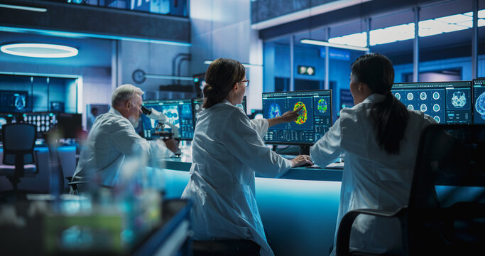 Back View Of Diverse Scientists Working In Medical Research Center. Male Doctor Using Microscope And His Female Colleagues Looking At MRI Brain Scans On Desktop Computers. Biotechnology Lab Concept.