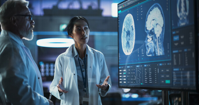 Neuroscience Laboratory Meeting: Female Asian Scientist And Male Caucasian Surgeon Discussing MRI Brain Scan On TV Display. Specialists Working On Medication For Rehabilitation After Brain Damage.