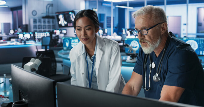 Asian Female Scientist Using Desktop Computer And Talking To Caucasian Male Doctor In Medical Hospital Research Lab. Colleagues Discussing Test Results Of Patients, Treatment Methods, Medication.