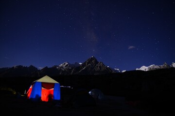 A tent of trekkers with light & shadows from its inside, while camping at the brink of Biafo glacier in Pakistan, with a partially visible milkyway on the moonlit top of mountain in the background. 