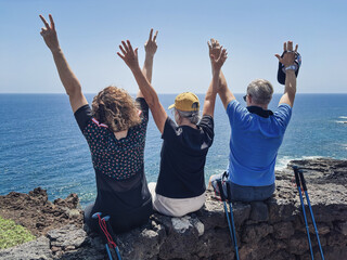 Back view of three friends in outdoor excursion at sea, sitting on the cliff looking at the horizon...