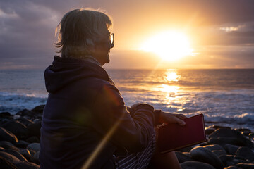 Senior smiling woman in vacation or retirement sitting on a pebble beach at sunset looking the...