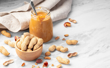 Peanuts with shell in wooden bowl with peanut butter in jar on white marble table