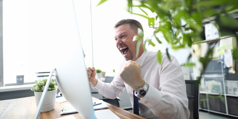 Portrait of cheerful man looking at computer monitor with gladness and making successful gesture....