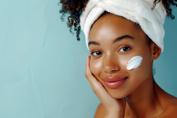 Young african american woman with towel on head applying cosmetic cream on face