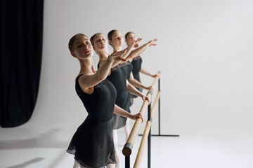 Ballet practice at barre. Beautiful young girl, ballet dancers in black costumes training, preparing for performance against grey studio background. Concept of ballet, art, dance studio, youth