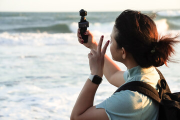 Young asian woman wearing backpack is taking a selfie at the beach using pocket gimbal camera with...