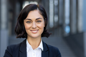 Close-up portrait of a beautiful young woman in a business suit standing outside a building, smiling and looking at the camera