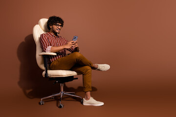 Full size photo of nice young man sit chair smart phone empty space wear shirt isolated on brown color background