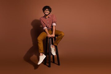Full size photo of nice young man sit stool empty space wear striped shirt isolated on brown color...