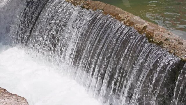 water flowing over concrete barrier in a row of thin trickles in small river dam at sunny day, closeup with slow motion