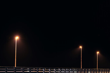 Street lampposts casts dim yellow warm along shore pier standing against pitch black canvas of...
