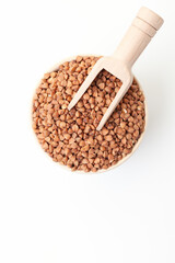 Dry, brown buckwheat grains in bowl with wooden scoop on white background. Top view. Space for a text