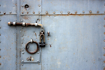 A fragment of an old gate, which is covered with iron. A rusty padlock hangs on an iron bolt. Ring handle on an old door with rivets on iron sheets. The door is locked. A rough lock on the door - obrazy, fototapety, plakaty