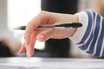Schoolgirl or student holding a pen in her hand - writing a dictation, preparing an answer at an exam or filling out documents in the classroom, sitting at a desk. Photo. Selective focus - obrazy, fototapety, plakaty