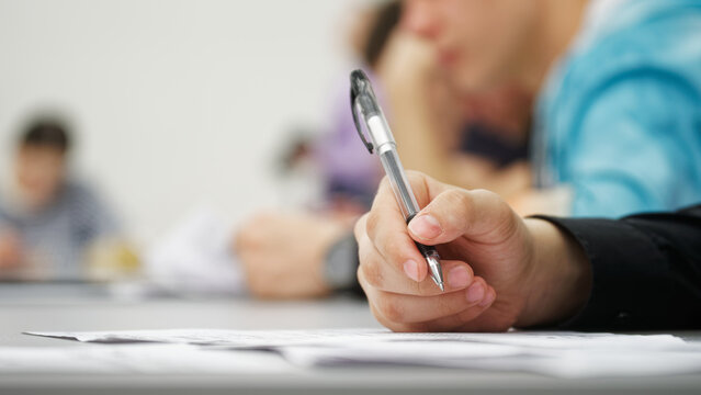 Guy student or schoolboy, writing a dictation, essay or filling out documents in the classroom, sitting at a desk next to other students. Photo. Selective focus. Close-up
