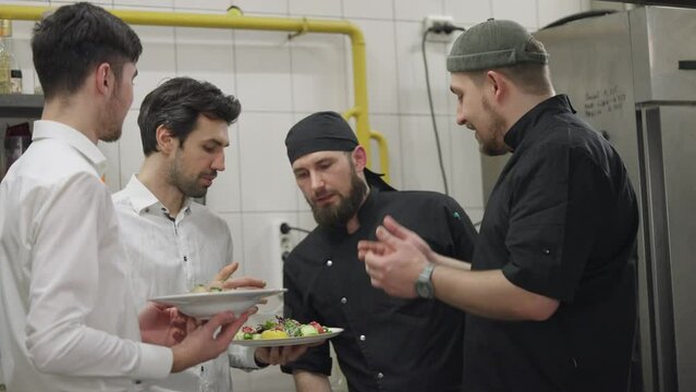 Two chefs in black uniforms together with waiters in white shirts communicate about the prepared dishes and solve their problems while working in the kitchen and restaurant. Cooks and waiters solve