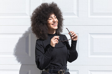 Young white woman with afro-style hair, dressed in black, leaning against a white door, smiling,...