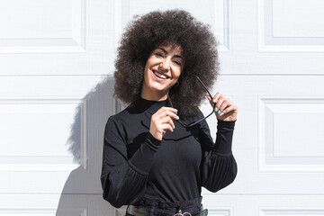 Young white woman with afro-style hair, dressed in black, leaning against a white door, smiling and...