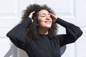 Young white woman with afro-style hair, dressed in black, leaning against a white door, smiling,...