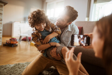Joyful father playing with son while mother takes photo with smartphone at home